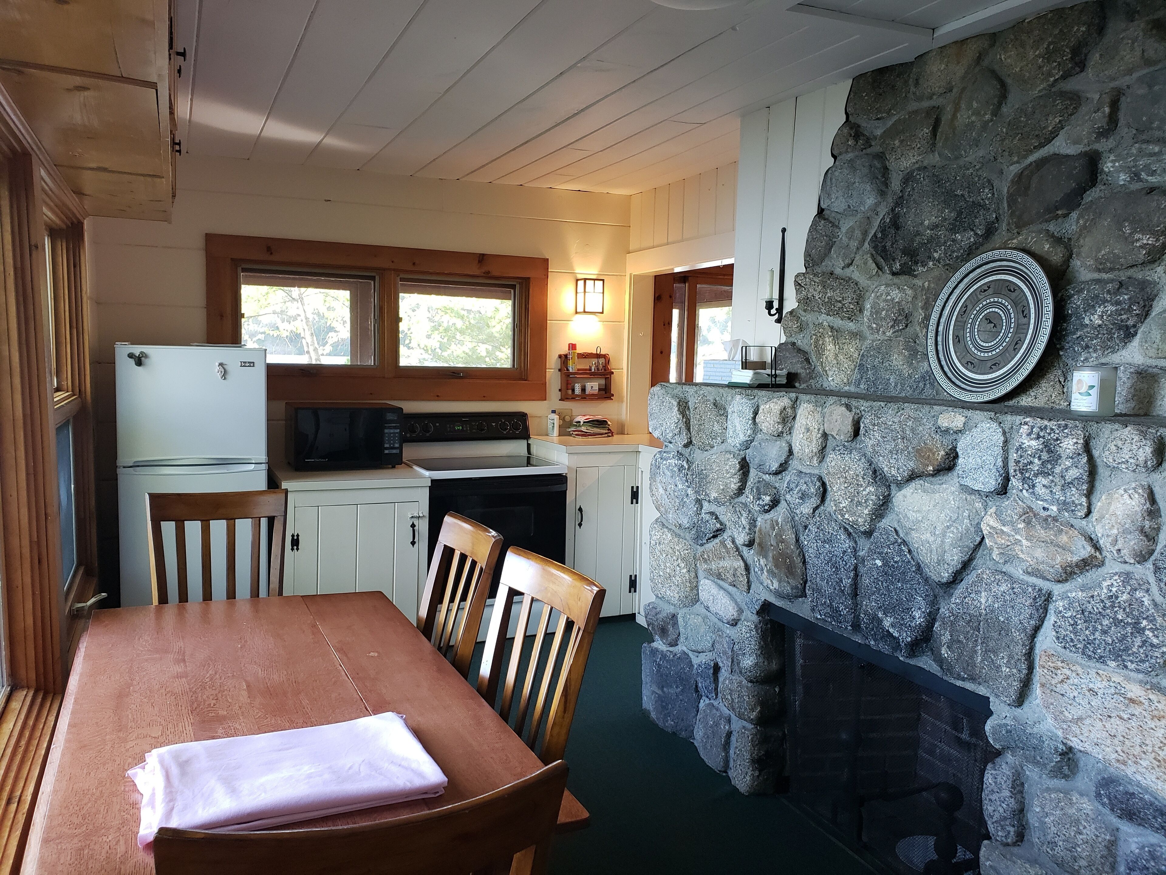A cozy cabin interior with a stone fireplace, wooden dining table, and a kitchen area with a refrigerator and microwave.