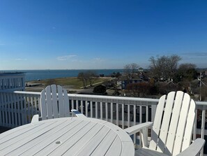 A deck with white Adirondack chairs and a table overlooking a coastal view with houses and a pier in the distance.