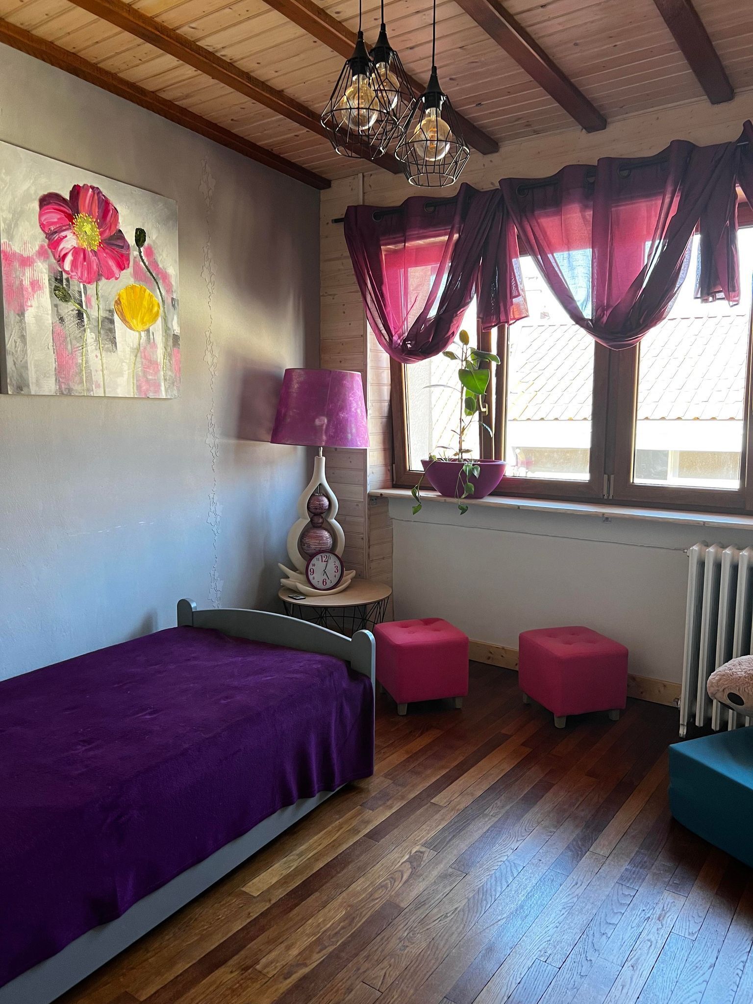 A bedroom with a bed, a nightstand, a lamp, a clock, a window with curtains, a radiator, and a plant.