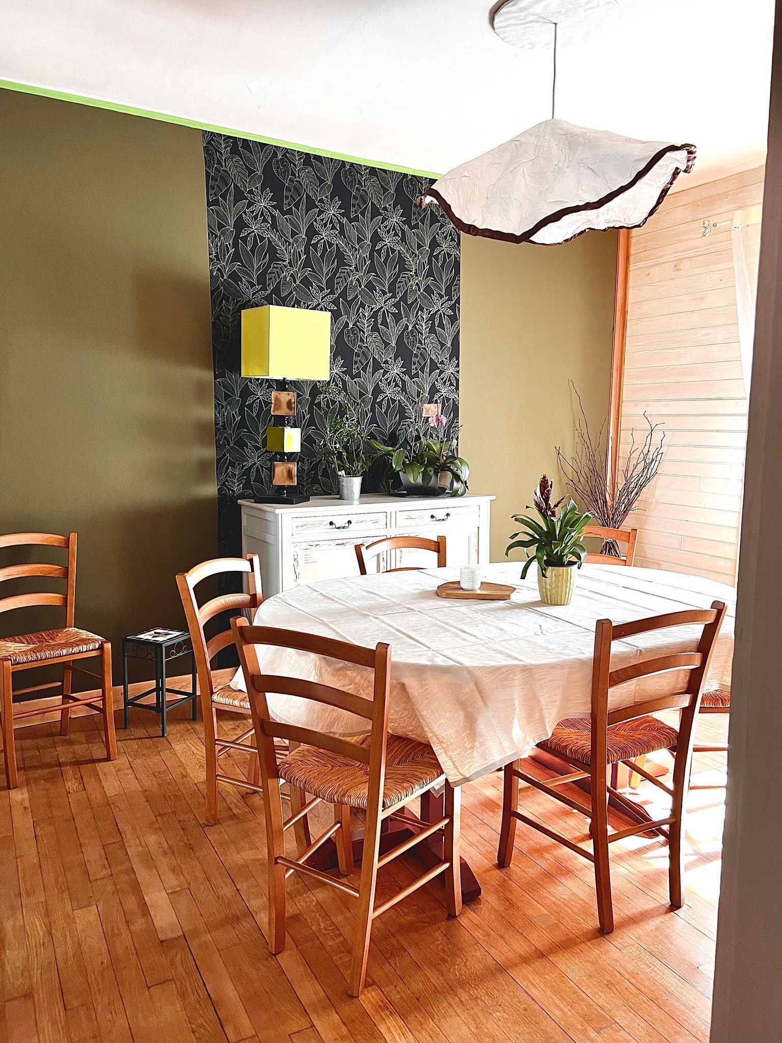 A dining area with a round table, wooden chairs, a white tablecloth, a green wall with a floral pattern, a yellow lamp, and a potted plant.