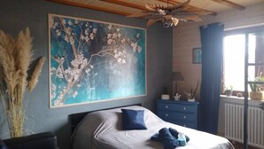 Una camera da letto con un grande quadro floreale, un letto con cuscino blu, una cassettiera e una finestra con piante.