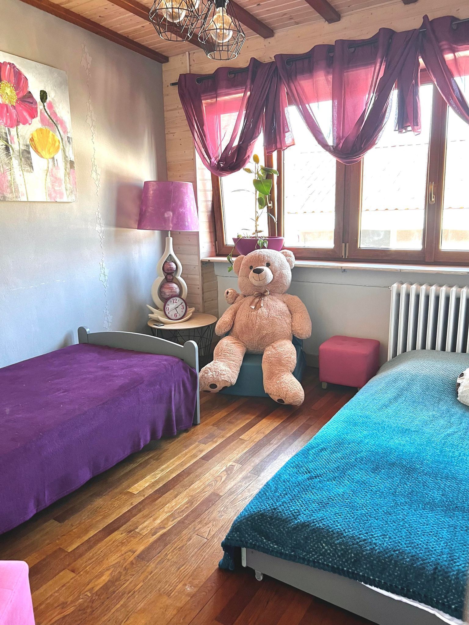 A room with two beds, a large teddy bear, a potted plant, a clock, and a window with curtains.
