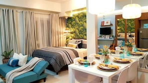 A modern bedroom with a bed, a sofa, a dining table set for two, and a kitchen area with a refrigerator and a television.