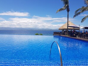 Une piscine d’un bleu éclatant, bordée d’une structure au toit de chaume et entourée de palmiers, offrant une vue sur une mer calme et des montagnes lointaines.