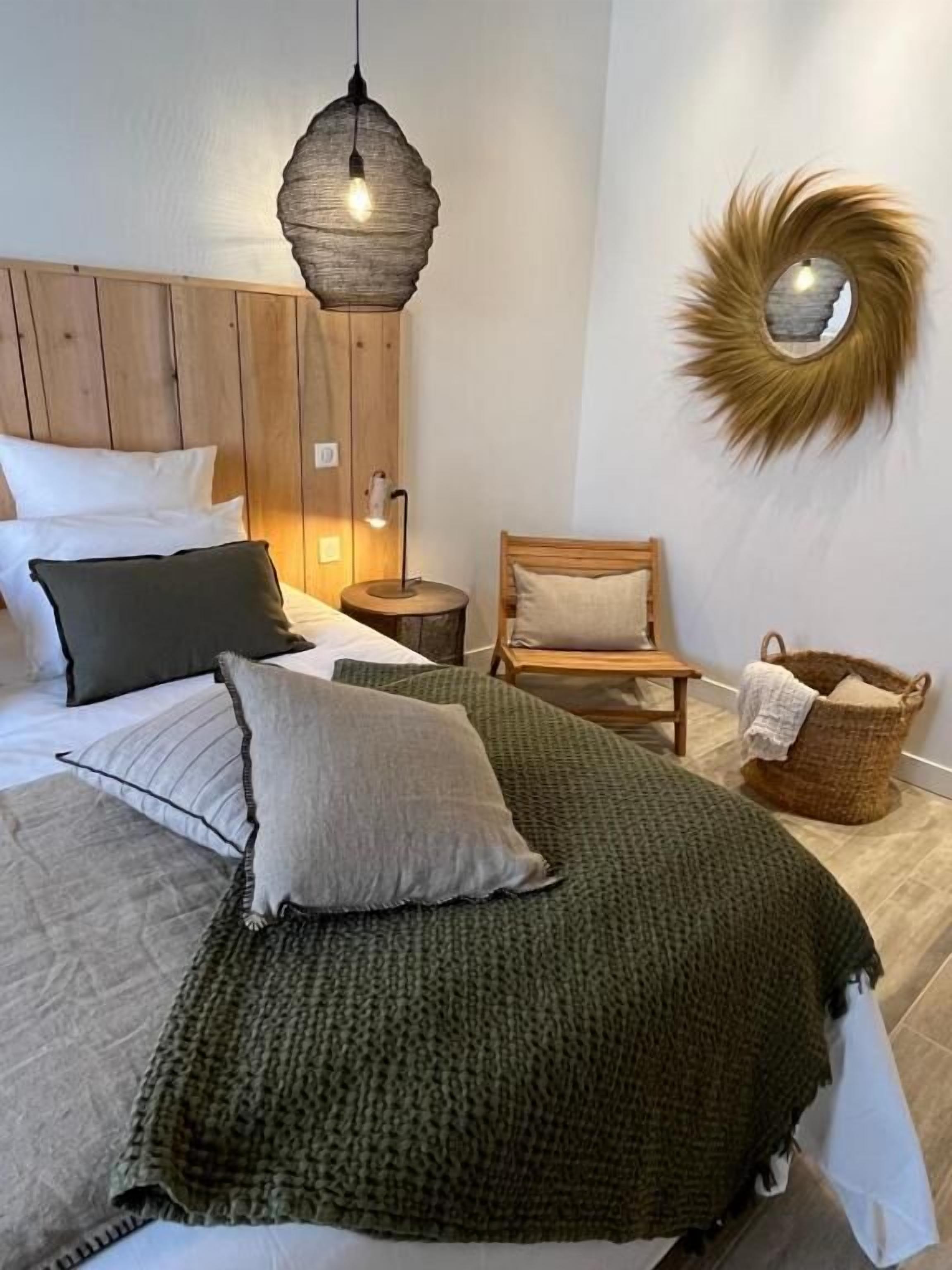 Un dormitorio con una cama grande, cabecera de madera, lámpara colgante, una mesita redonda con una lámpara, una silla y un espejo decorativo en la pared.