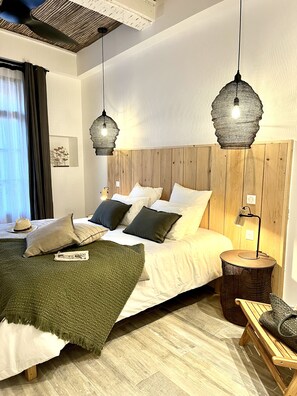 Un dormitorio con cabecera de madera, cama con ropa blanca, dos lámparas colgantes, una lámpara de noche y una mesita redonda con lámpara.