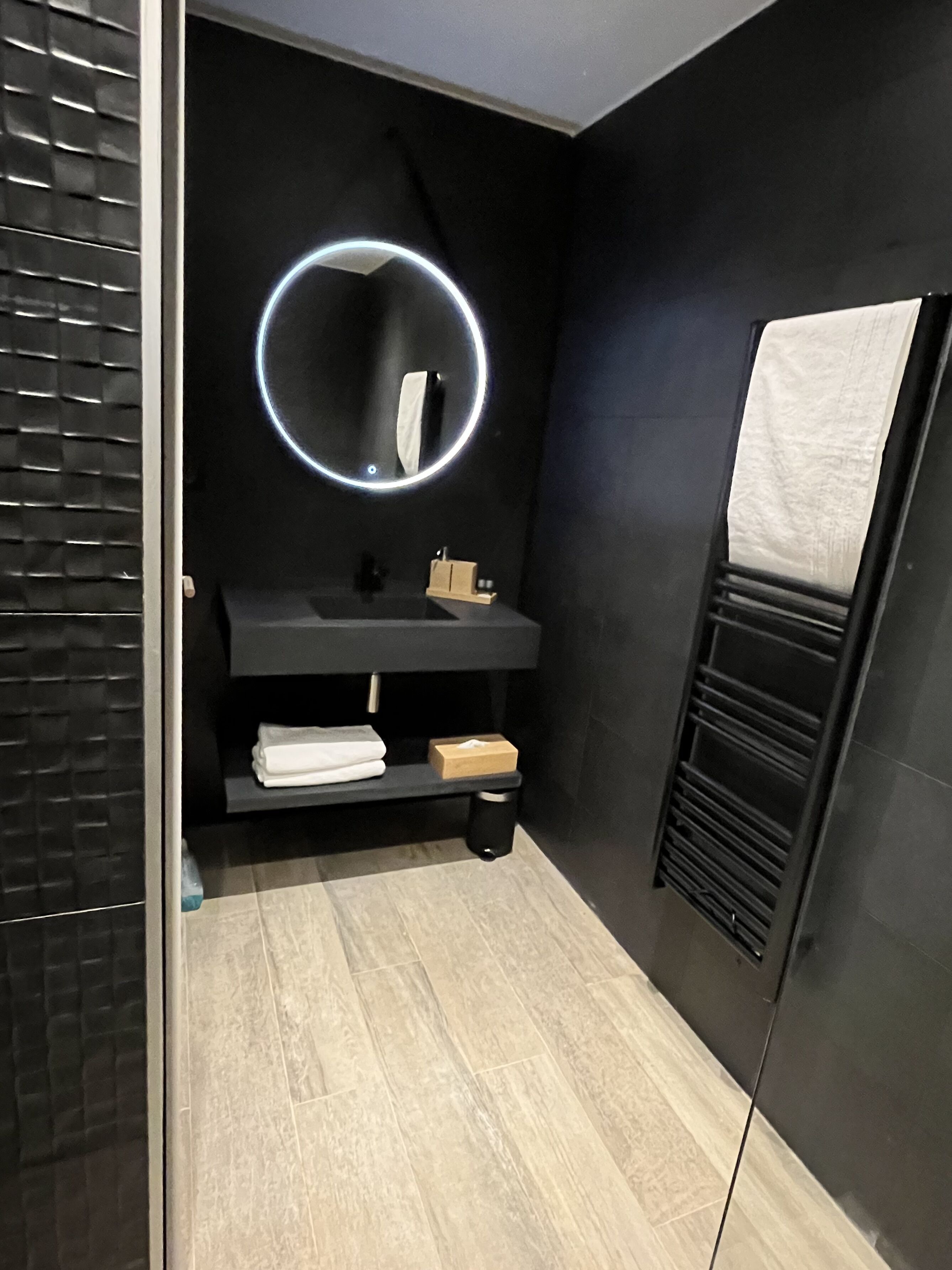 Un baño moderno con un tocador negro, un espejo redondo con luz, un perchero y una toalla en la pared.