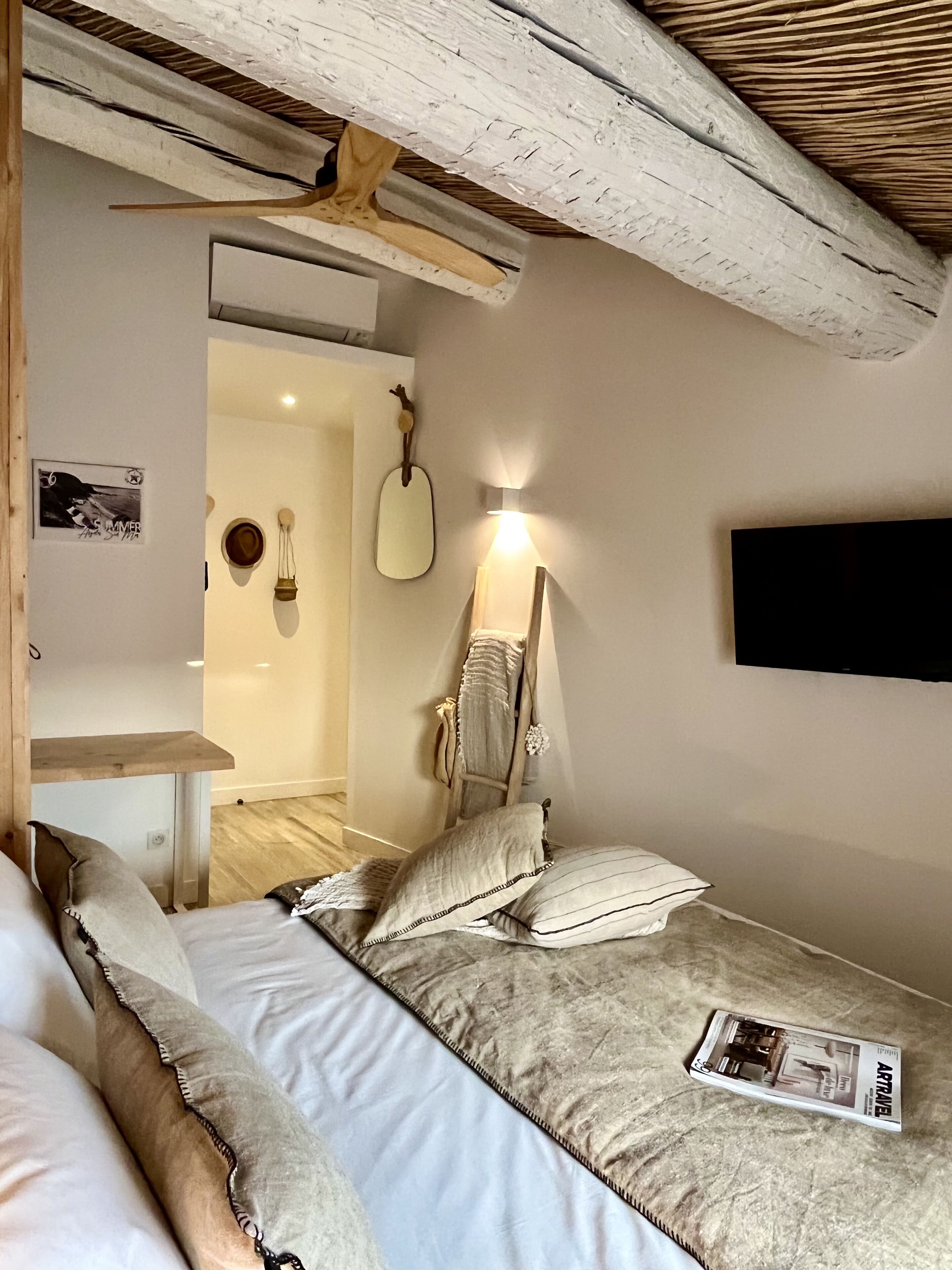 Un dormitorio con cama, almohadas, una mesita de noche, un televisor montado en la pared y una viga de techo de madera.
