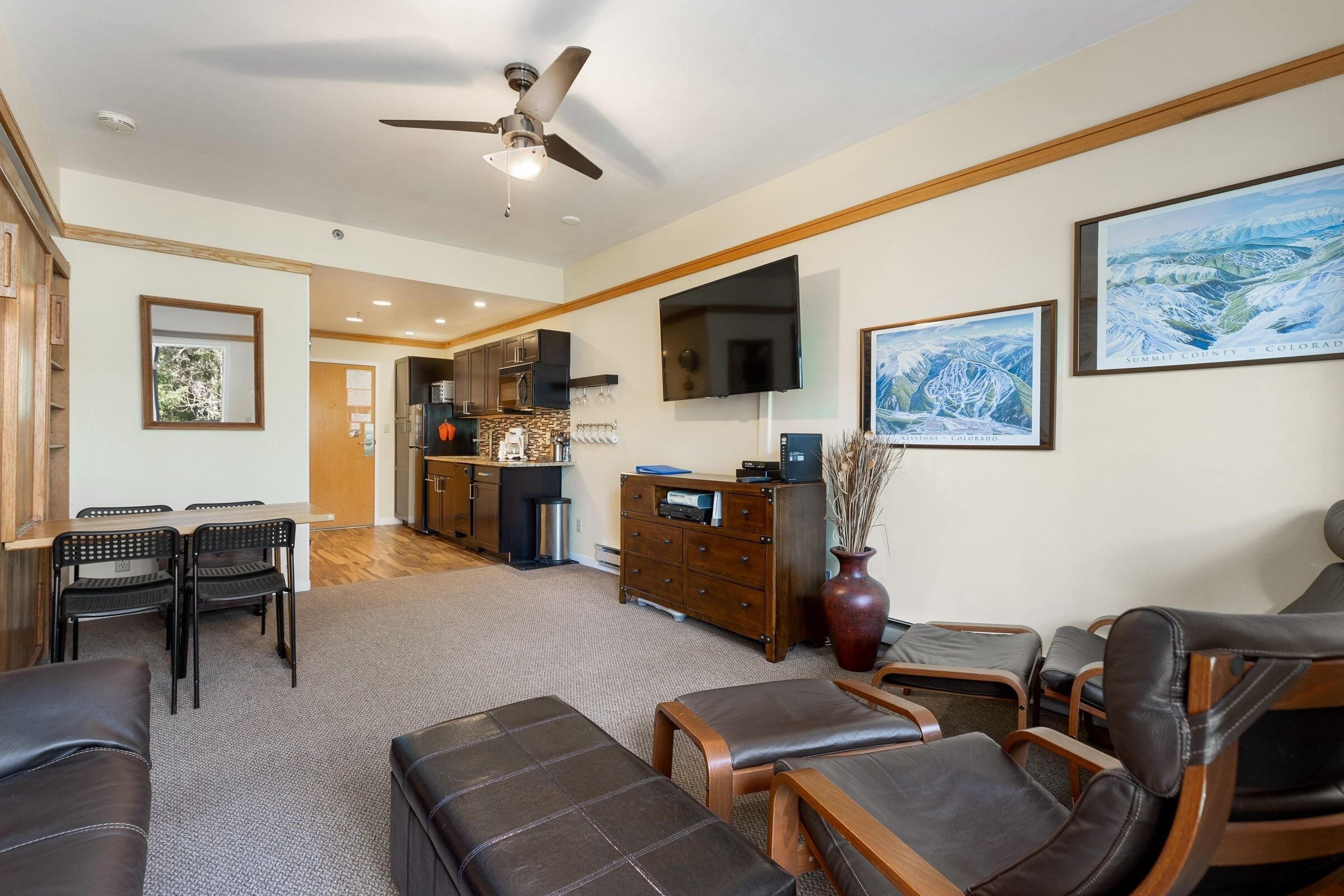 A spacious living room with a ceiling fan, a dining area with a table and chairs, a kitchen in the background, and a large TV mounted on the wall.