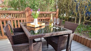 A wicker outdoor dining set with a glass table and chairs, a bottle of wine, and two wine glasses on a deck.