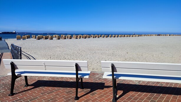 A beach with a row of wooden bollards, two white benches with black armrests, and a clear blue sky.