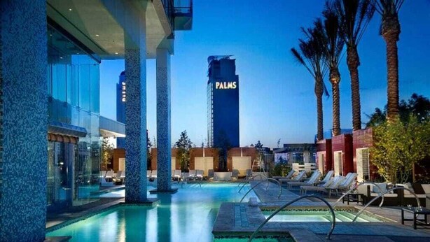 Ein Hotelpoolbereich mit Liegestühlen, Palmen und einem hohen Gebäude im Hintergrund mit der Aufschrift 'PALMS'.