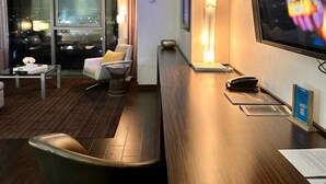 Ein modernes Hotelzimmer mit großem Fernseher, einer Couch, einem Schreibtisch und Blick auf die beleuchtete Stadt.