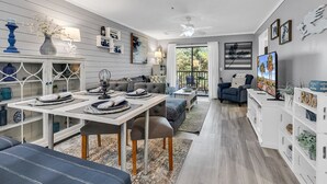A modern living room with a dining area, a sofa, a TV, and a bookshelf. - Charming 2 Bed 2 Bath True Blue Pawleys Island Golf, 4 Pools Beach 5 min drive! (Pawleys Island)