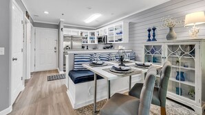 Una cocina moderna con muebles blancos, un comedor con una mesa dispuesta para cuatro personas y un banco empotrado con cojines azules.