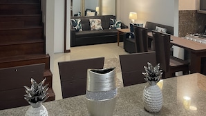Una sala moderna con una escalera de madera oscura, un sofá negro con cojines tropicales, un comedor con mesa de granito y una cocina con estufa a gas.