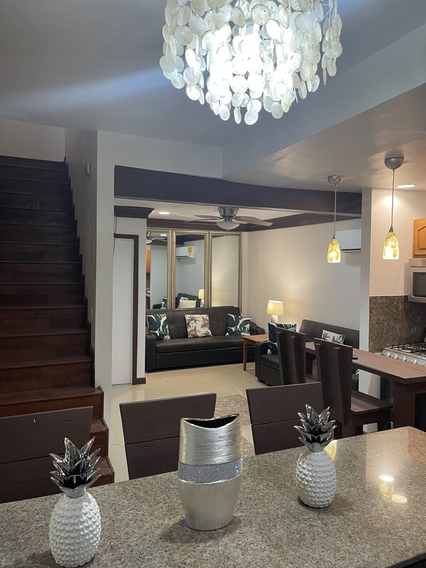 Un salon moderne avec un escalier en bois foncé, un canapé noir agrémenté de coussins aux motifs tropicaux, un coin repas avec une table en granit et une cuisine équipée d’une cuisinière à gaz.
