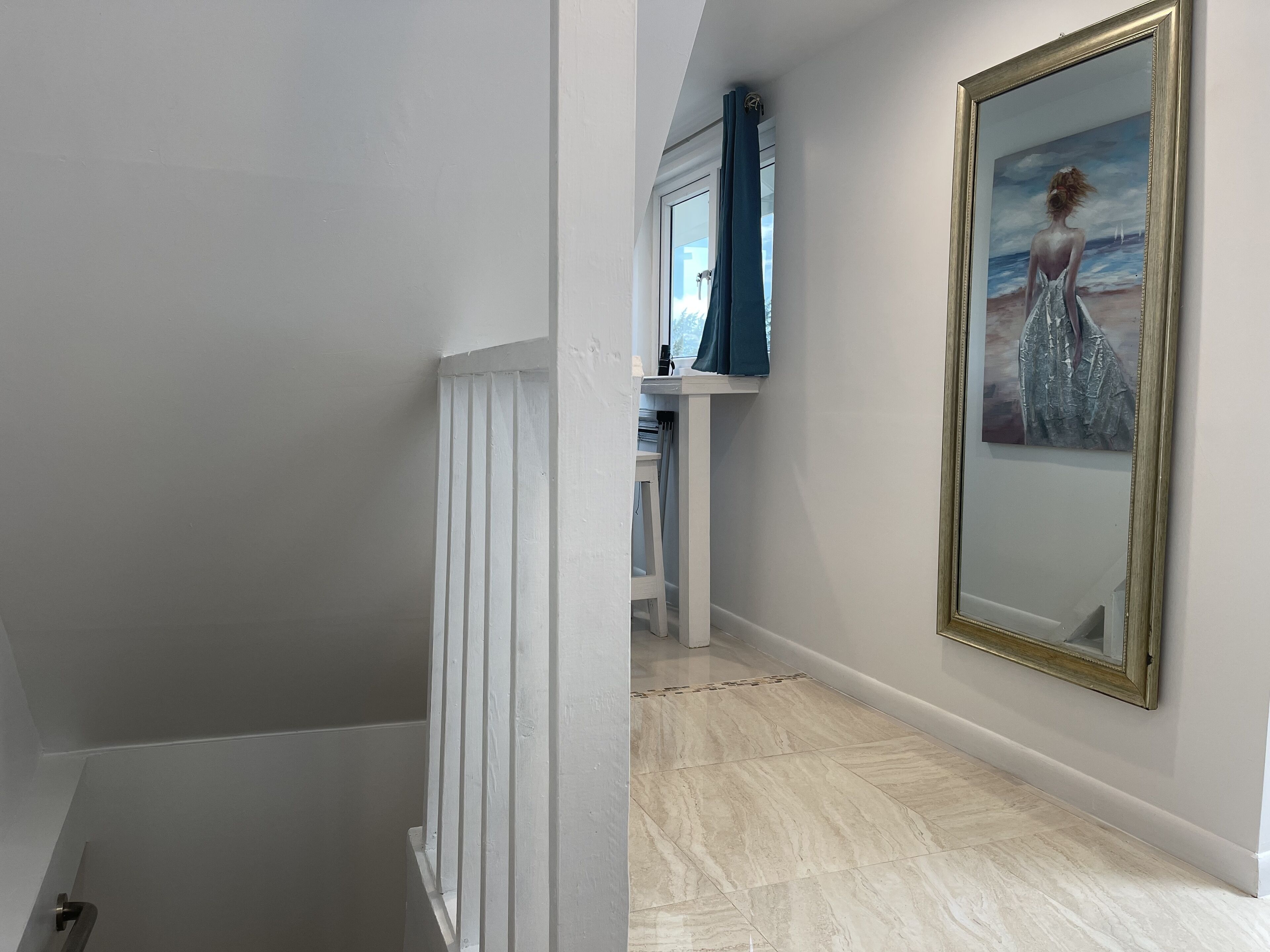 Um corredor com corrimão branco, um quadro de uma mulher na parede e uma janela com cortinas azuis.