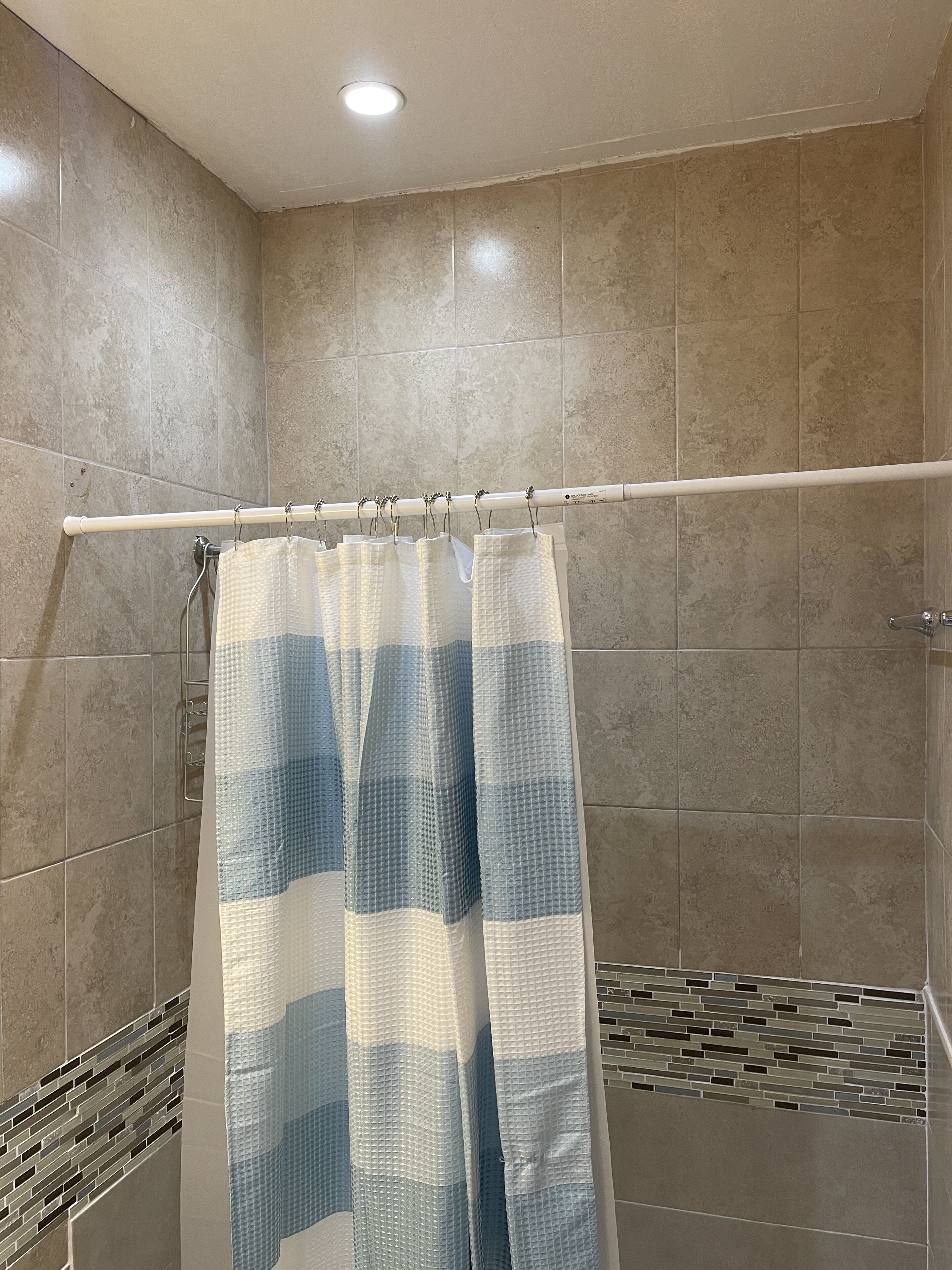 Um chuveiro com cortina de chuveiro quadricula branca e azul, paredes com azulejos bege e uma luminária branca no teto.
