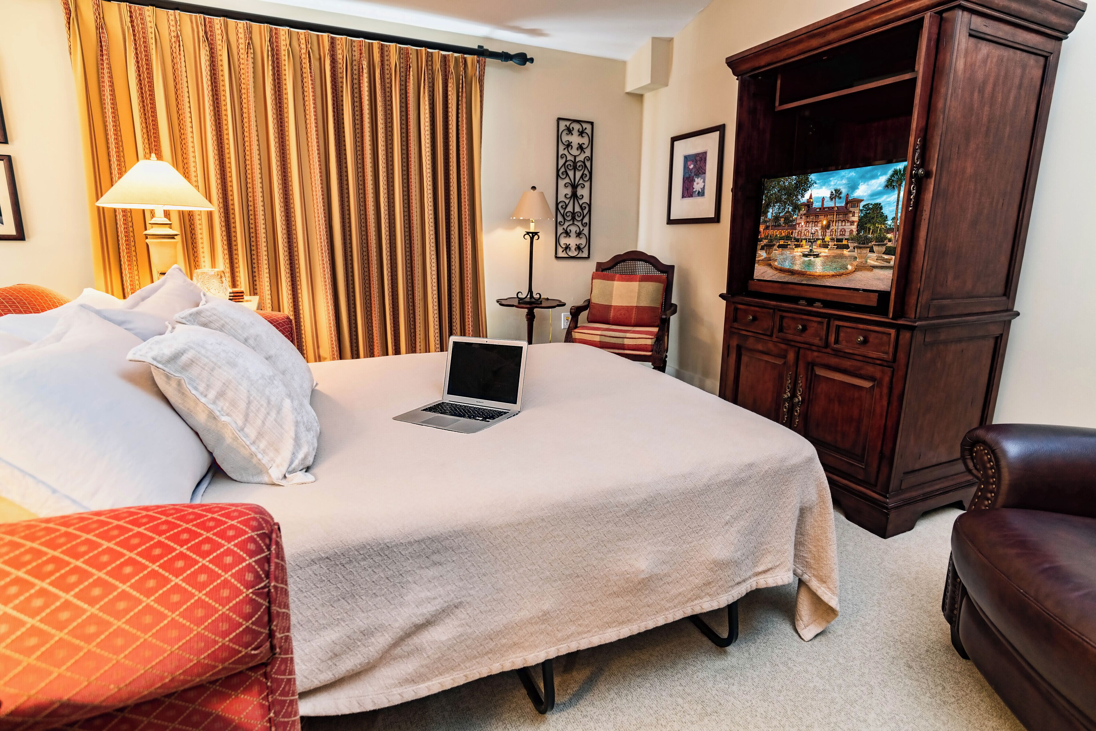 A hotel room with a bed, a laptop, a TV, a chair, and a lamp.