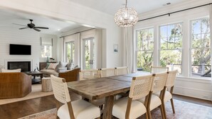 A spacious living room with a large wooden dining table, comfortable seating, a ceiling fan, and large windows offering a view of the outdoors.