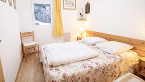 Ein Schlafzimmer mit einem hölzernen Bett, einem Sessel, einem Fenster mit Blick auf Bäume und einem kleinen Regal mit einer Vase.