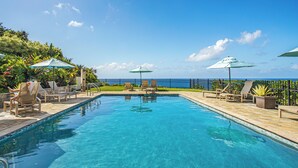 A clear blue pool with lounge chairs and umbrellas, a view of the ocean, and a stone deck.