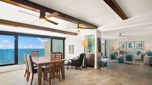 A spacious living area with a large dining table, chairs, and a view of the ocean. There are ceiling fans, a kitchen area, and a seating area with cushions.