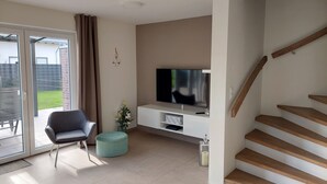 A modern living room with a flat-screen TV, a grey chair, a teal ottoman, and a staircase. - NEW! modern, high-quality, central apartment, sauna, garden, wallbox in Burhave (Butjadingen)