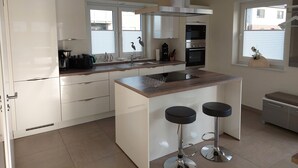 A modern kitchen with white cabinets, a stainless steel range hood, a double oven, and a window with blinds. - NEW! modern, high-quality, central apartment, sauna, garden, wallbox in Burhave (Butjadingen)