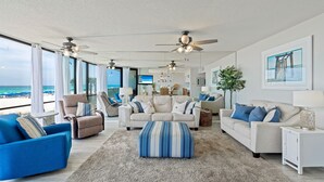 Uma sala de estar ampla com uma grande janela com vista para o oceano, um sofá bege, uma poltrona azul e um banquinho listrado.