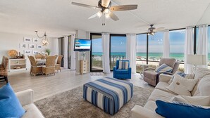 Uma sala de estar espaçosa com uma grande janela com vista para a praia, um ventilador de teto e uma área de jantar com móveis de vime.