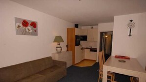 A compact living space with a brown sofa, a dining table with chairs, a kitchen area with cabinets, and a wall-mounted clock.