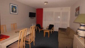A small, furnished room with a dining table, chairs, a sofa, a television, and a window with curtains.