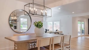 A modern dining area with a wooden table, four chairs, a large round mirror, and a hanging light fixture. - @ Marbella Lane - Cozy Modern Home w/ Relaxing Design & Pool (Fullerton)