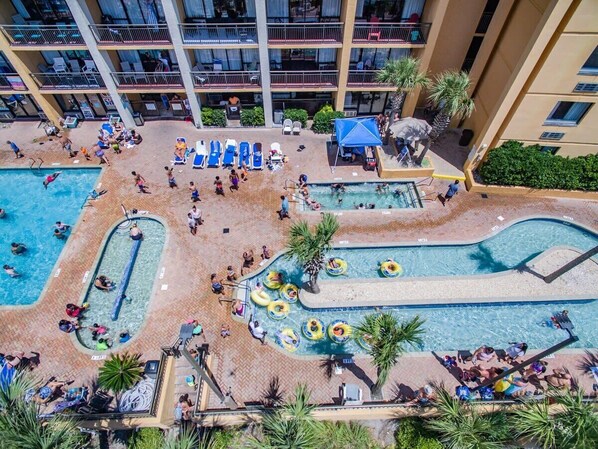 An aerial view of a hotel pool area with multiple swimming pools, lounge chairs, and palm trees.