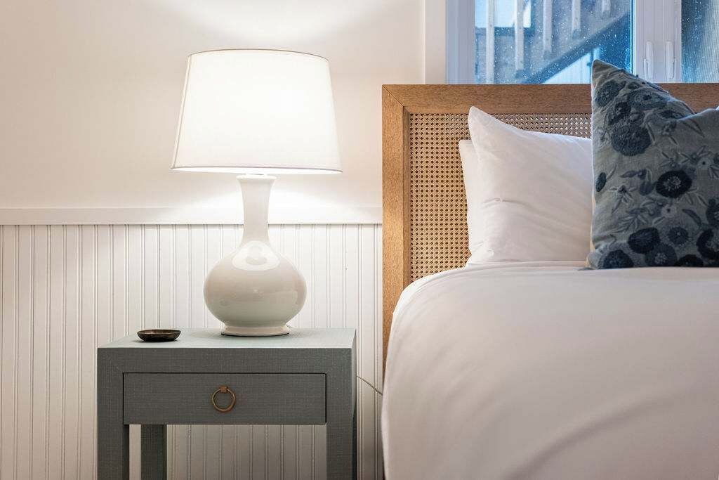 A neatly made bed with a white comforter and a blue floral pillow, a white bedside lamp on a small wooden nightstand with a drawer, and a window with a view of a building.