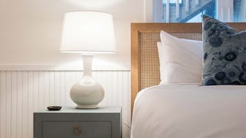 A neatly made bed with a white comforter and a blue floral pillow, a white bedside lamp on a small wooden nightstand with a drawer, and a window with a view of a building.