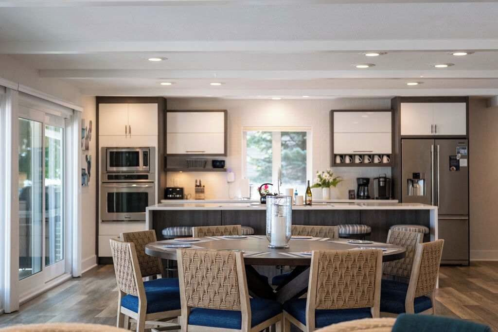 A modern kitchen with built-in appliances, a dining area with round tables and patterned chairs, and a large window providing natural light.