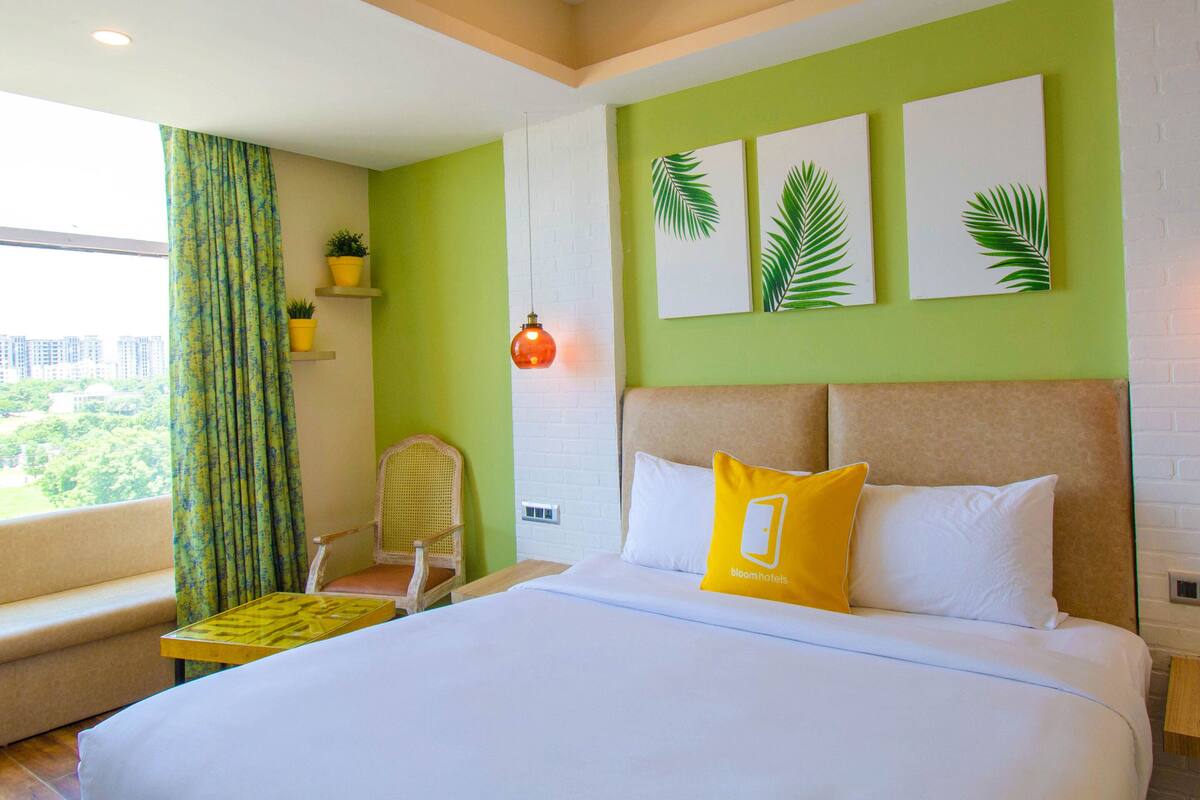 Habitación de hotel con una cama grande, una silla, un ventanal con cortinas y una pared decorada con tres cuadros de hojas de palmera.