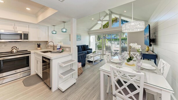 A modern kitchen with white cabinetry, a built-in oven, and a microwave. There is a dining area with a glass table and white chairs. - Coconuts101-BEACH FRONT COMPLEX-1BR ground level-ByIslandRealEstate (Holmes Beach)