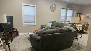 Ein Wohnzimmer mit einem dunkelbraunen Sofa, einem Fernseher auf einem Holzboden, einem Esstisch mit Stühlen und einer Wanduhr.