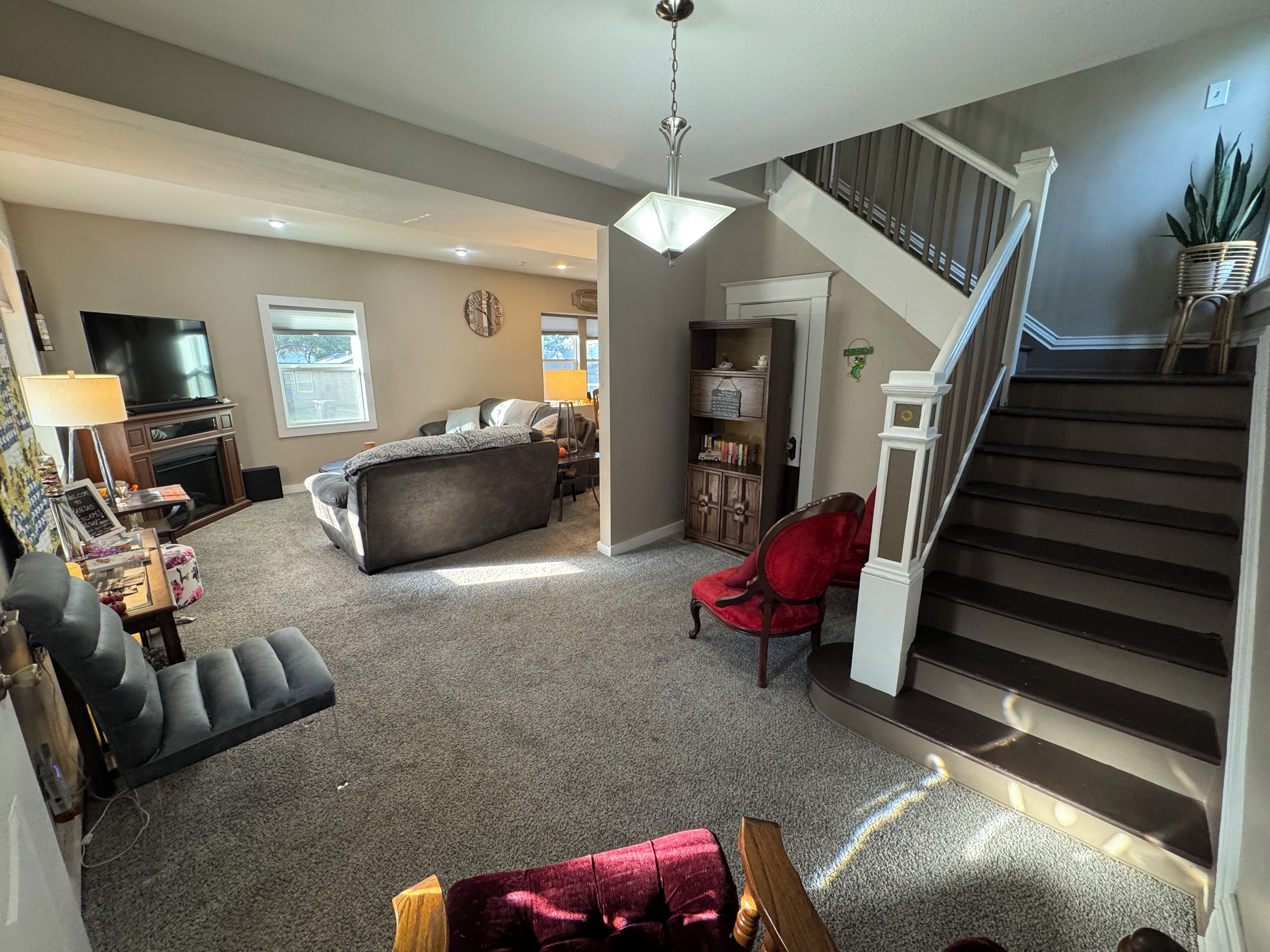 Een ruime woonkamer met een open haard, een grote televisie, een comfortabele bank en een rode fauteuil. Een trap leidt naar een verdieping.