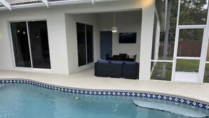A swimming pool with a tiled edge, a dark-colored outdoor seating area, and a television mounted on the wall. - Retreat at Genoa  (Port St. Lucie)