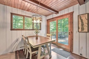 Un espace repas comprenant une table et des chaises en bois, un luminaire suspendu et de grandes fenêtres donnant sur la forêt.