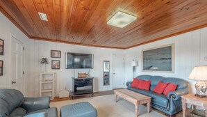 Un salon chaleureux avec un plafond en bois, un téléviseur à écran plat, un canapé bleu agrémenté de coussins rouges et une table basse.
