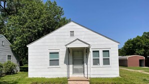 A small, single-story white building with a front door and windows, surrounded by grass and trees.