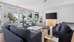 A modern living room with a ceiling fan, large windows, and a flat-screen TV. - Newly Built 7-bedroom Oceanfront Stunner with private pool. (Hilton Head Island)
