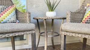 Una silla de mimbre con un cojín, una mesita redonda con una planta en maceta y otra silla con un cojín.