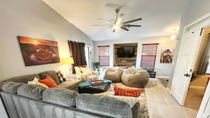 A living room with a grey sofa, a coffee table, and two bean bag chairs. There is a ceiling fan, a TV, and a door leading to another room. - Pool & EV Charger @ Family Friendly comfy house in the heart of Queen Creek (Queen Creek)